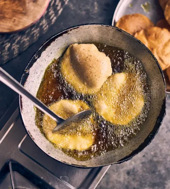 Puri frying in bubbling oil in a karahi on the stove, with a slotted spoon and finished breads alongside