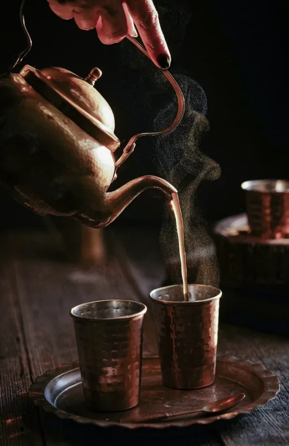 Steaming chai poured from a hammered copper kettle into copper cups on a wooden table